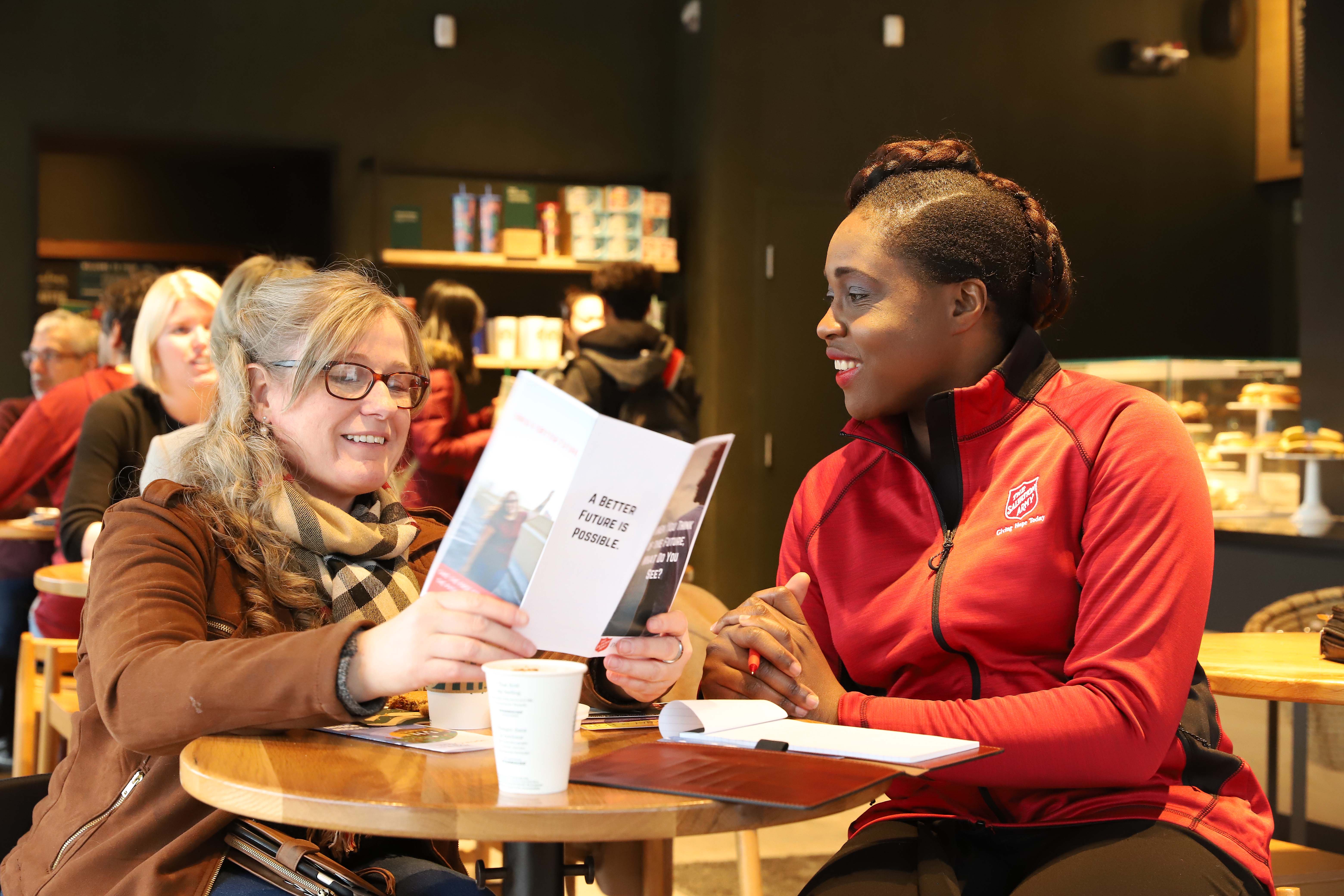 Staff member in a Salvation Army uniform sitting with a woman at a table, reviewing a booklet together in a welcoming community space.