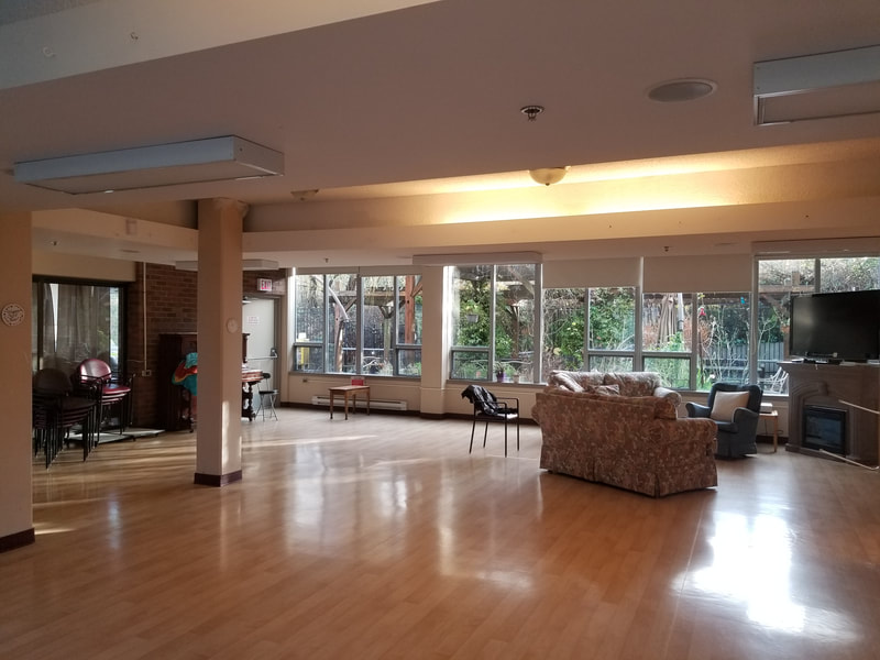 Spacious multi-purpose Sunset Room with hardwood floors, large windows overlooking greenery, seating areas, and open space for activities and events.